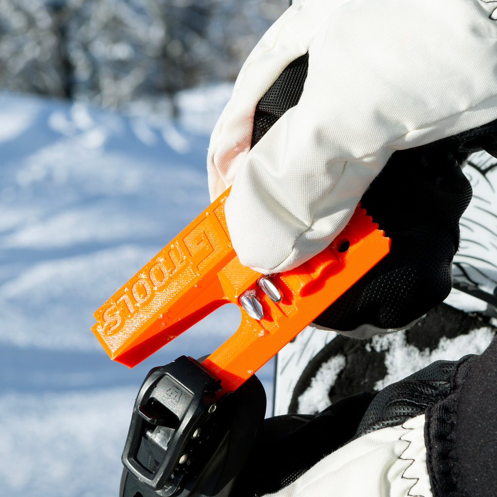 NG Snowboard Binding Tool – orange multitool on snow, used for cleaning snowboard ratchet buckles. Includes screwdriver, two bits, and a whistle. Perfect for tuning bindings, base, edges, and waxing.