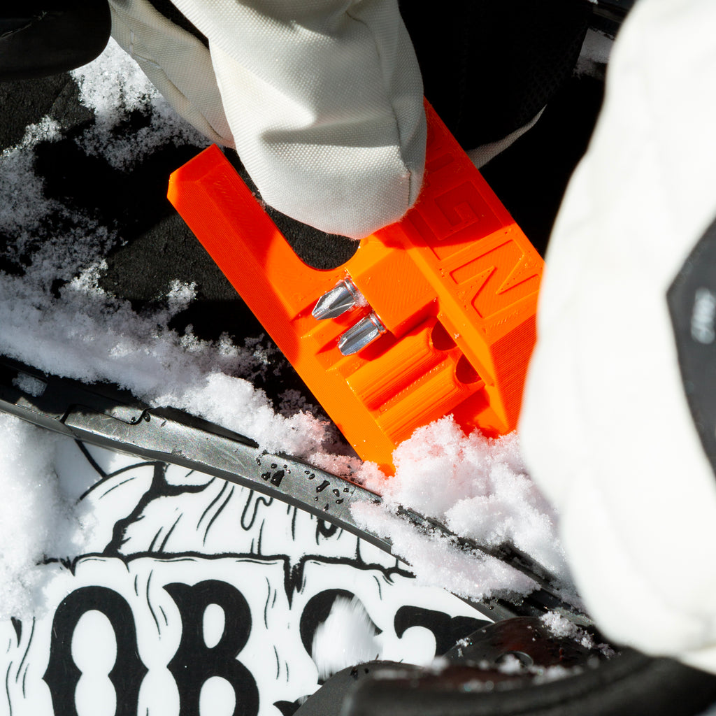 NG Snowboard Binding Tool – orange multitool on snow, cleaning ratchet buckles and removing snow from bindings. Includes screwdriver, two bits, and a whistle. Ideal for tuning bindings, base, edges, and waxing.