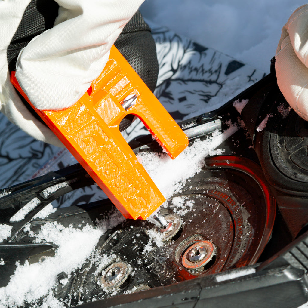 NG Snowboard Binding Tool – orange multitool on snow, cleaning ratchet buckles with a screwdriver. Includes two bits and a whistle. Perfect for tuning bindings, base, edges, and waxing.