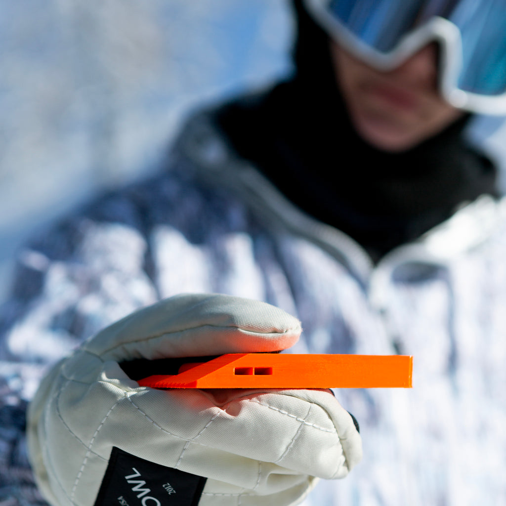 NG Snowboard Binding Tool – orange multitool on snow, cleaning ratchet buckles and using the whistle for a safety call. Includes screwdriver, two bits, and a whistle. Perfect for tuning bindings, base, edges, and waxing.