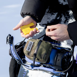 NGTOOLS Camo Snowboard Binding Bag attached to a snowboard binding high-back, compact and lightweight for secure storage. Paired with a yellow NGTOOLS small multitool for snowboard tuning.
